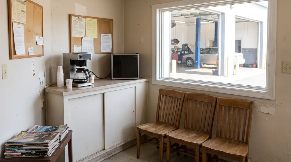 Sala de espera sencilla del taller: un mostrador, tres sillas, una cafetera y una ventana que da al taller